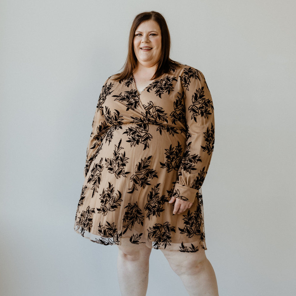 A person smiles while wearing a beige faux wrap dress with black floral patterns and black shoes, standing against a plain background.