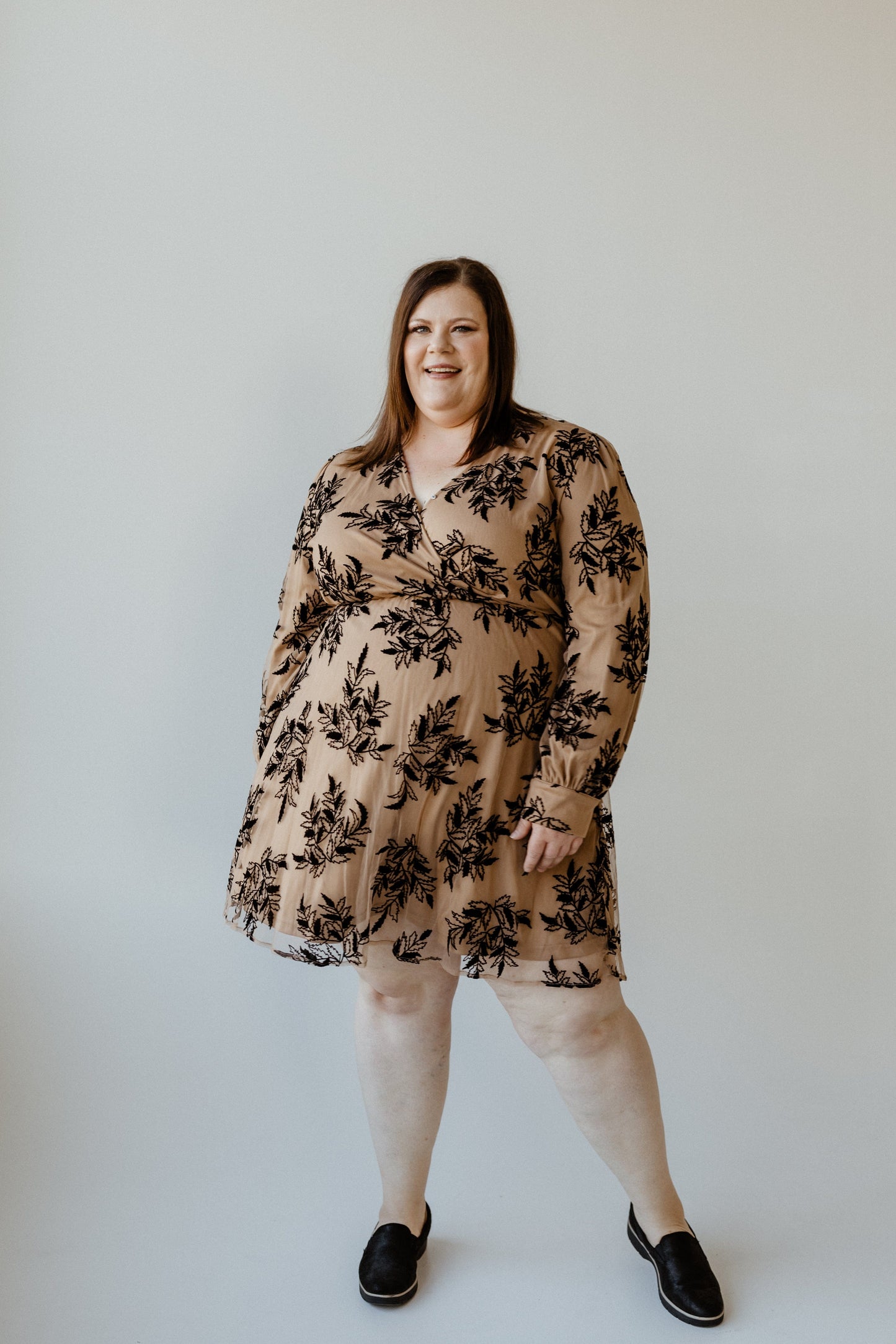 A person smiles while wearing a beige faux wrap dress with black floral patterns and black shoes, standing against a plain background.