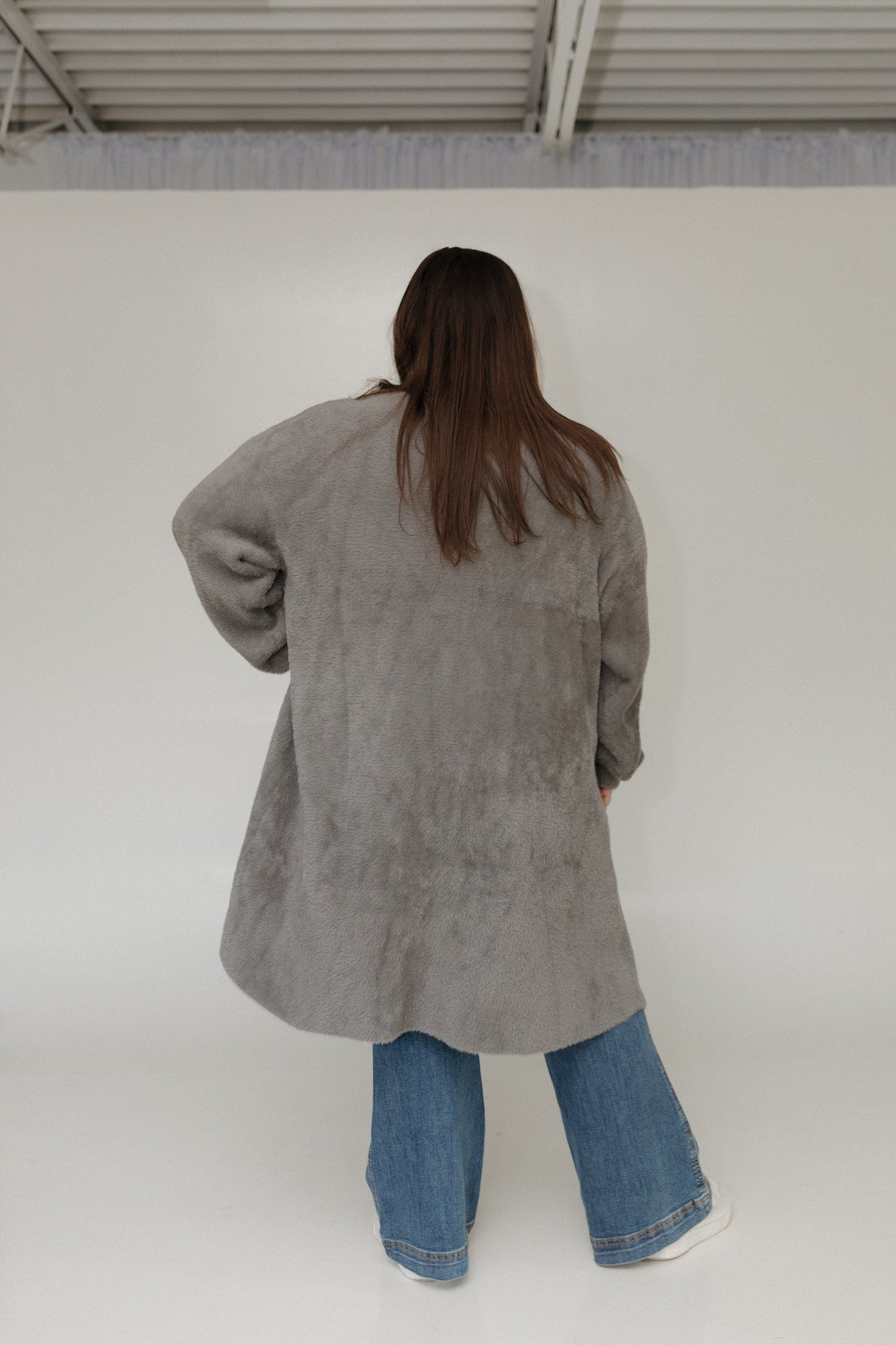 Person wearing a gray coat and blue jeans standing against a white wall.