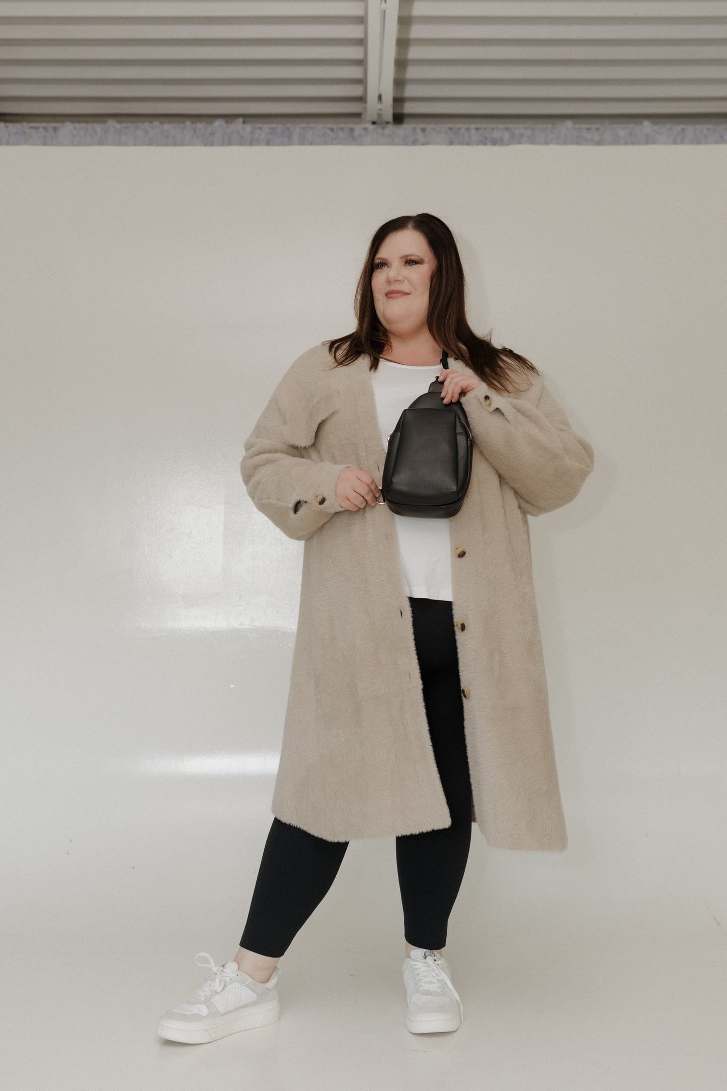 Woman wearing a beige trench coat holding a black handbag against a white background