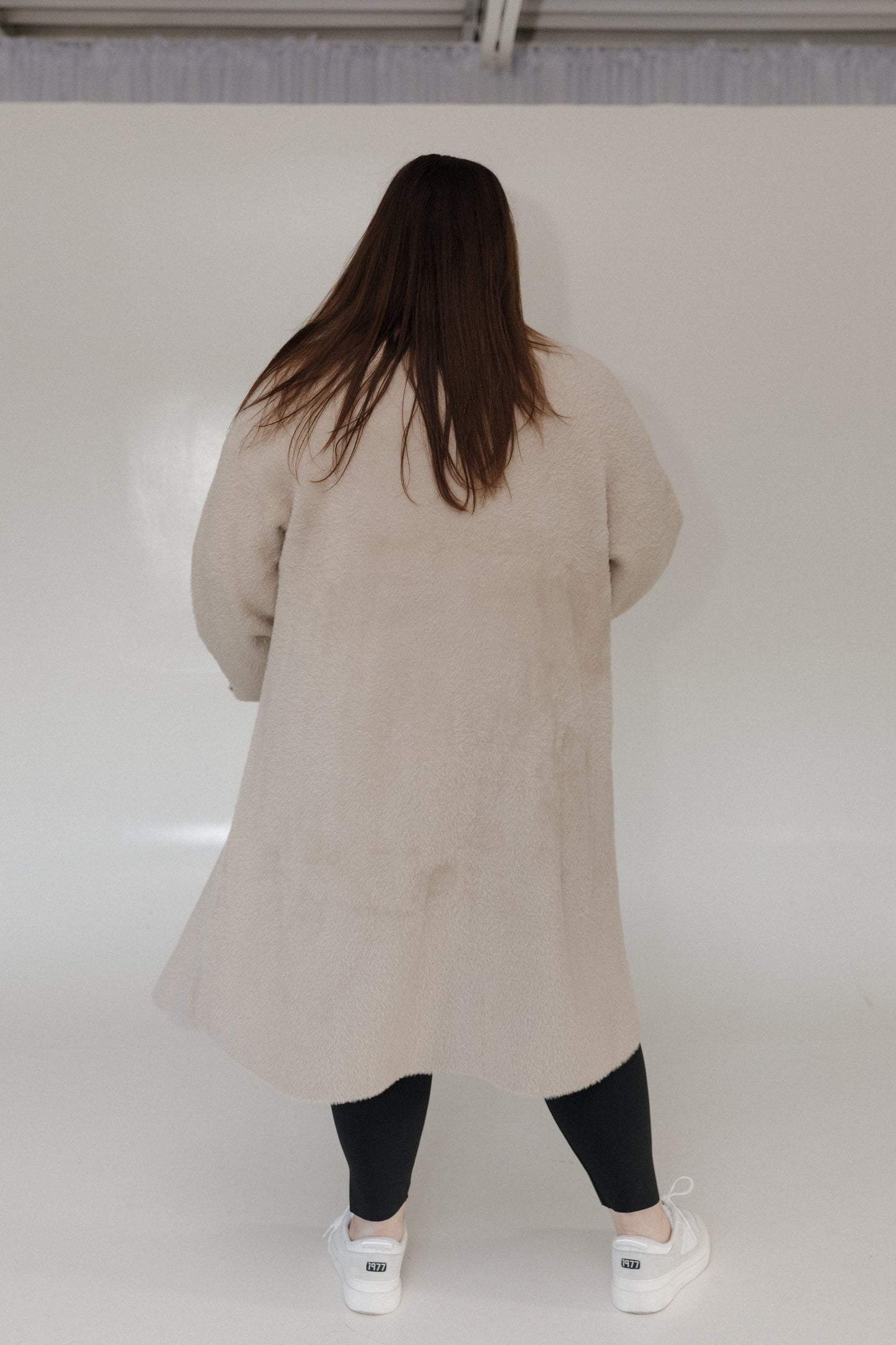 Person wearing a beige coat standing against a white wall