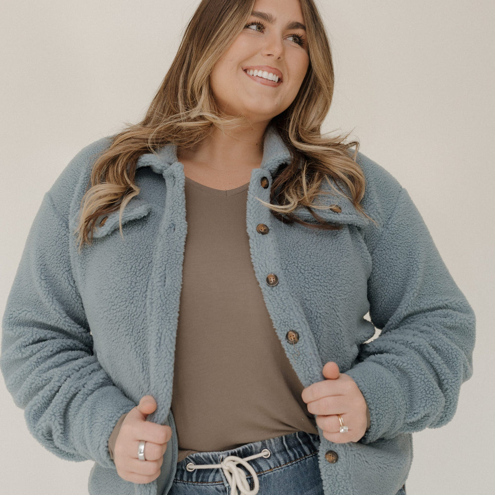 Woman wearing a light blue jacket over a brown shirt and jeans, standing against a plain background.