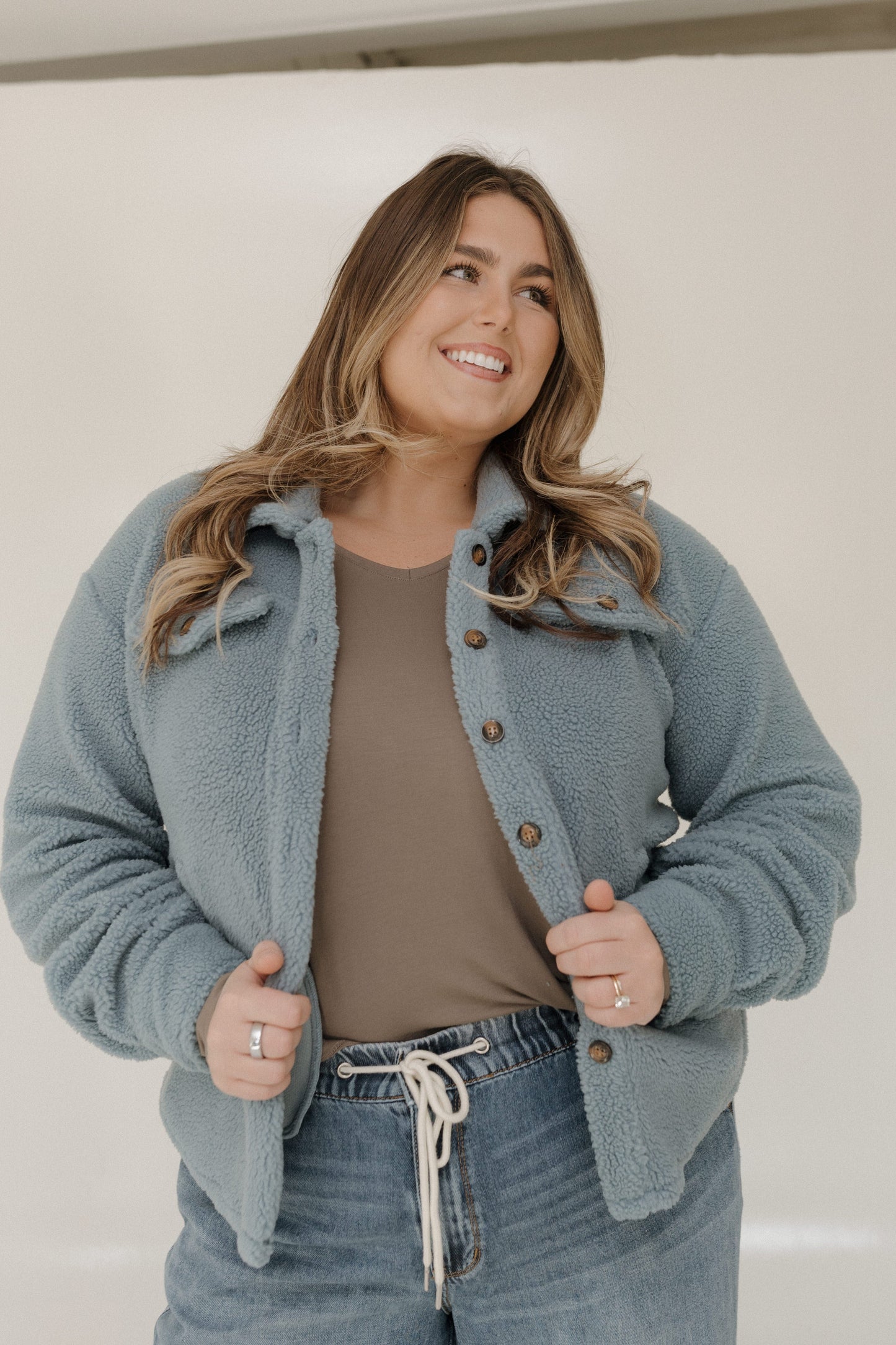 Woman wearing a light blue jacket over a brown shirt and jeans, standing against a plain background.