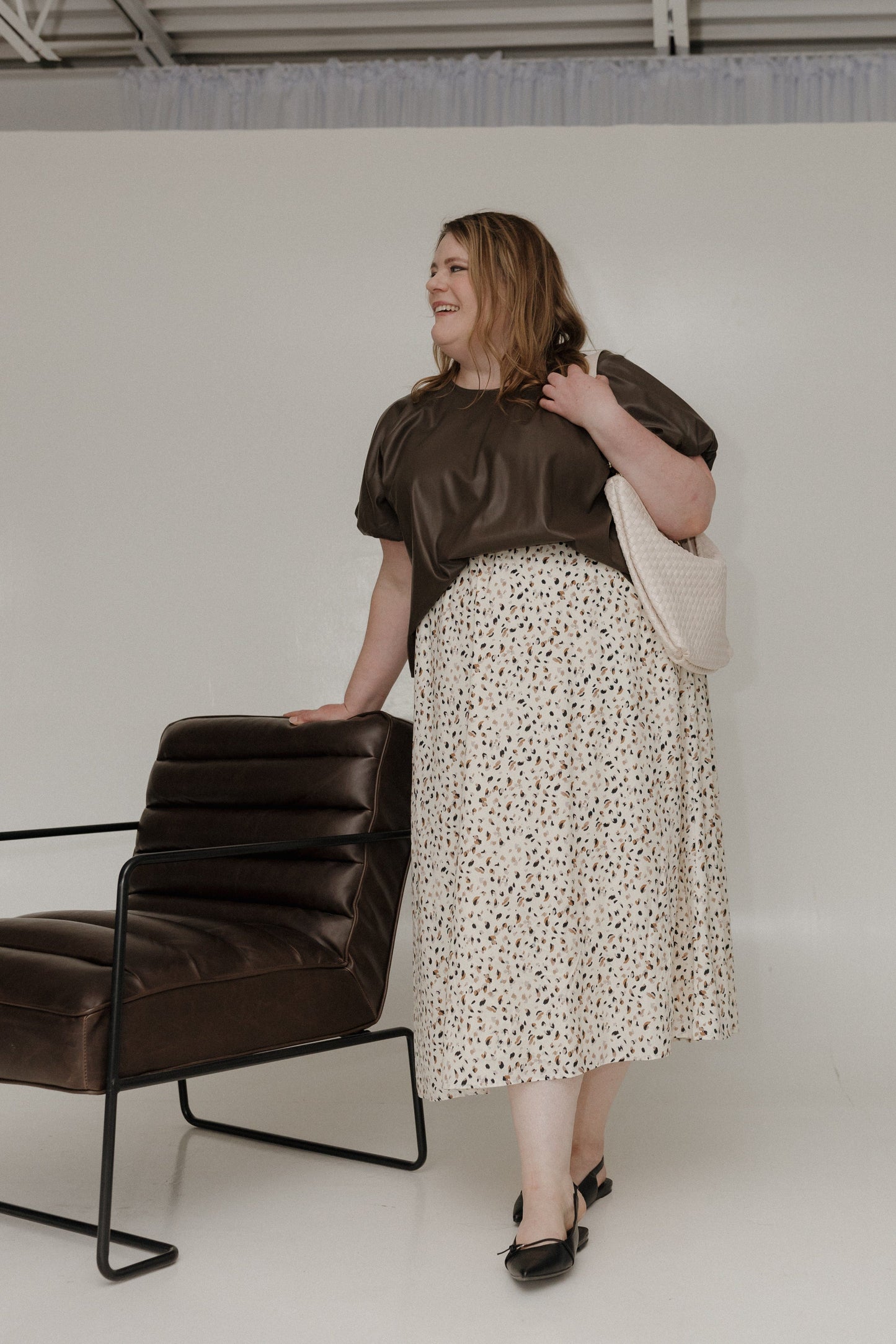 Woman in a brown top and patterned skirt standing next to a black chair against a white wall.