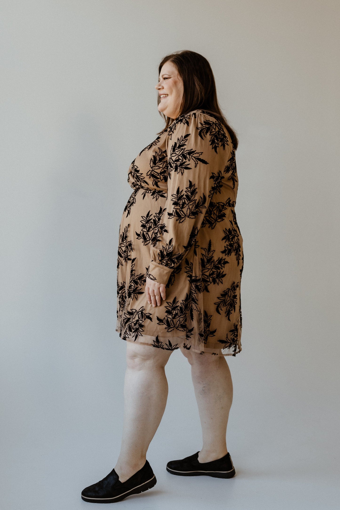 A woman stands in profile wearing a faux wrap dress in beige, adorned with black floral patterns and complemented by her black slip-on shoes.