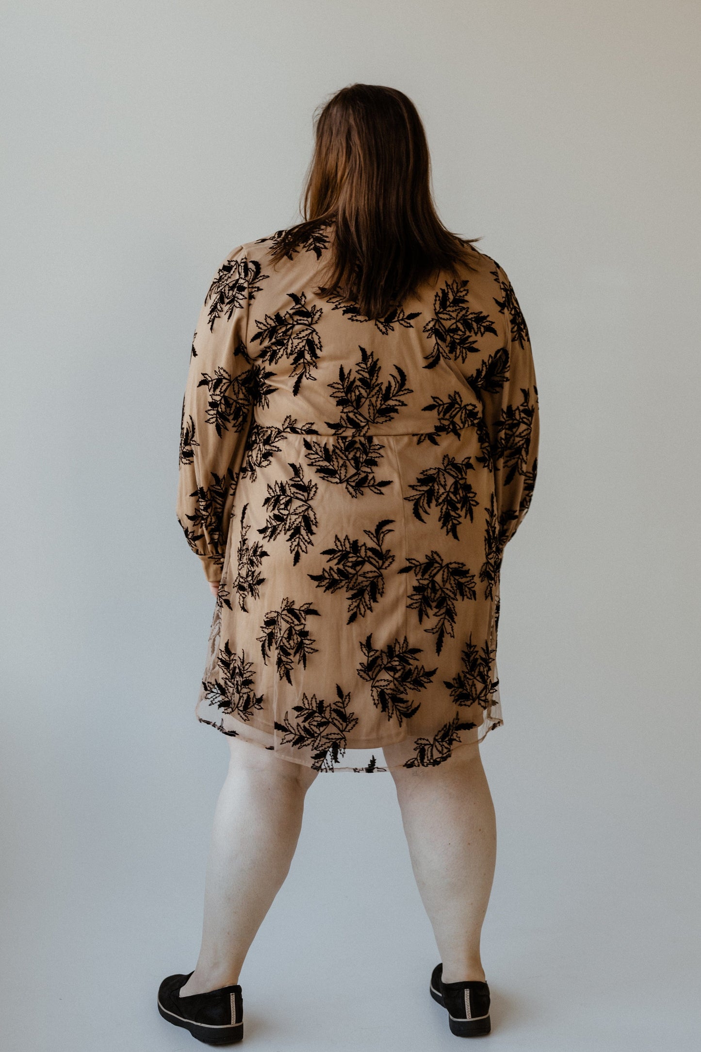A person with long hair stands with their back to the camera, wearing a beige faux wrap dress adorned with black floral patterns and black shoes.