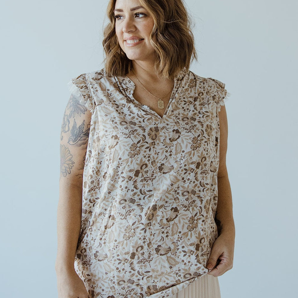 A woman with shoulder-length hair, wearing the FEMININE CAP SLEEVE BLOUSE IN SAND by Love Marlow and a pleated skirt, smiles while looking to the left. Her right arm displays visible tattoos, exuding an air of sophisticated fashion.