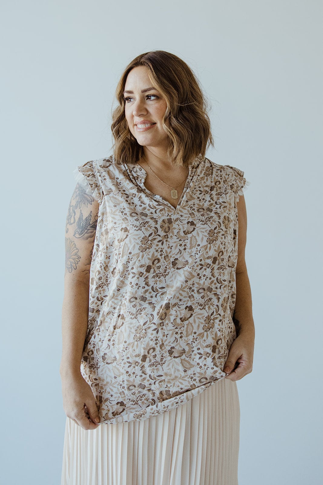 A woman with shoulder-length hair, wearing the FEMININE CAP SLEEVE BLOUSE IN SAND by Love Marlow and a pleated skirt, smiles while looking to the left. Her right arm displays visible tattoos, exuding an air of sophisticated fashion.