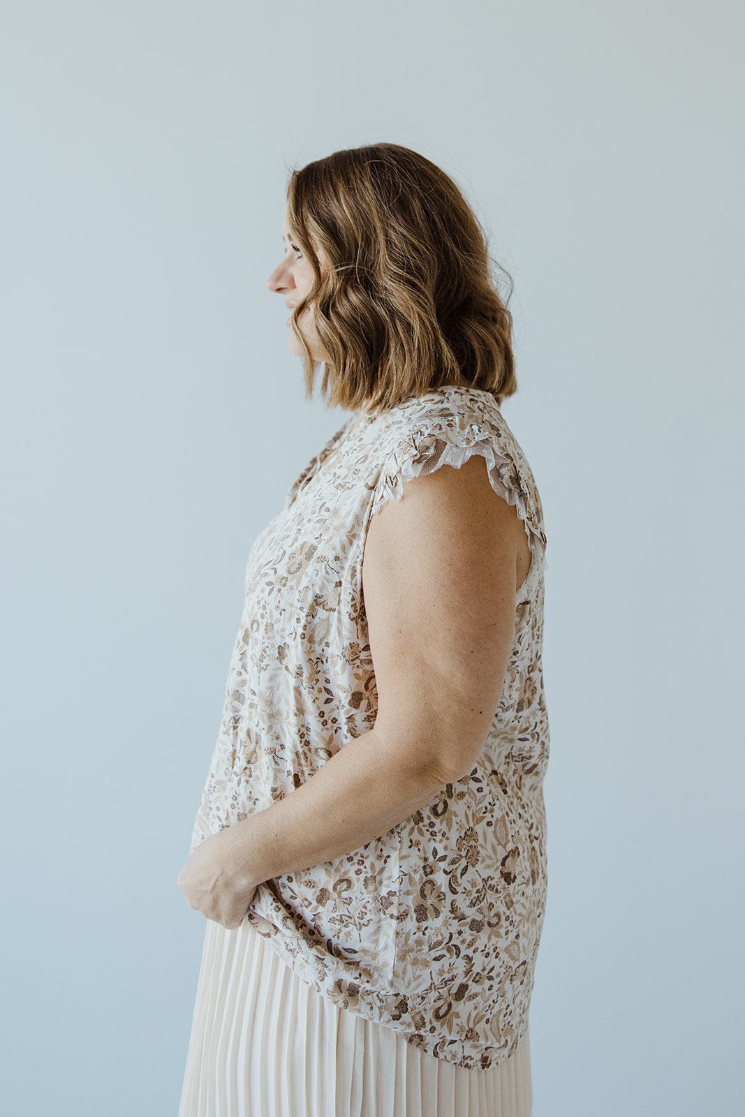 A woman with shoulder-length hair in a sophisticated, Love Marlow FEMININE CAP SLEEVE BLOUSE IN SAND and pleated skirt stands in profile against a plain background.