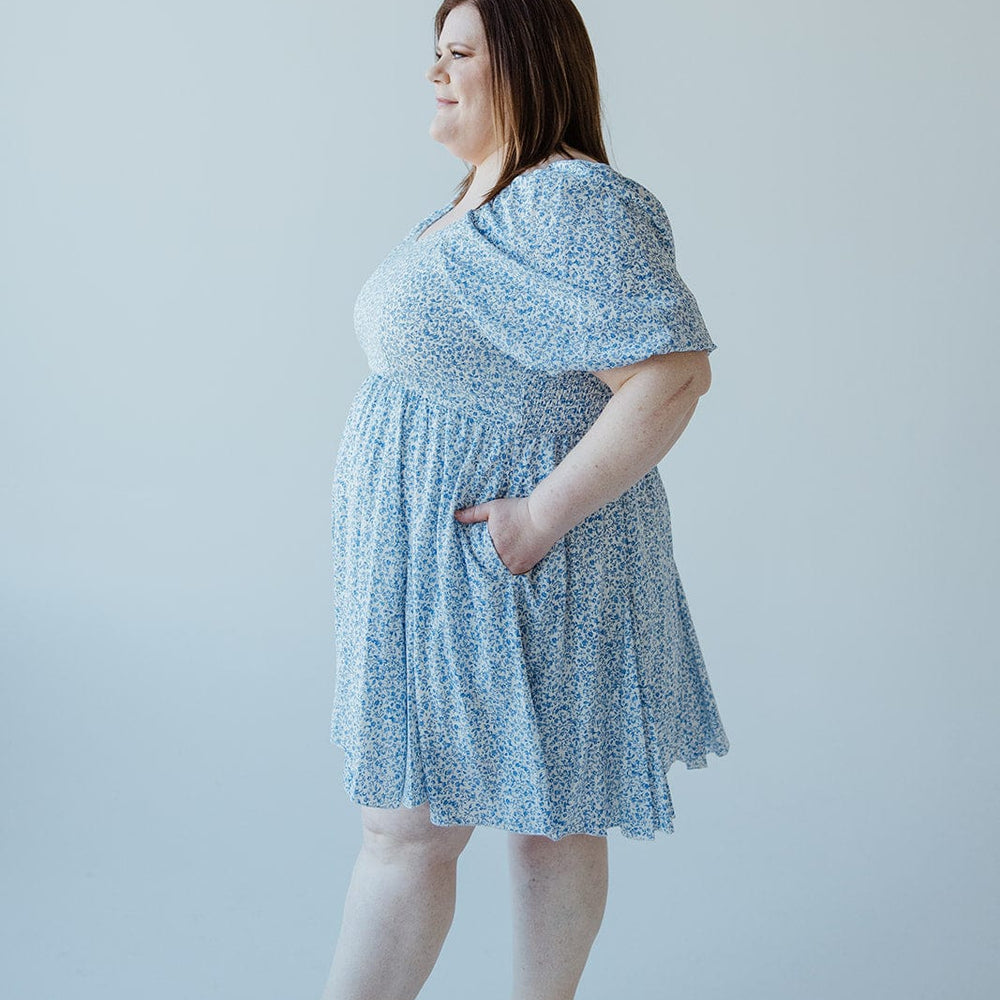 A woman is standing in profile, wearing a light blue FEMININE FLORAL PUFF SLEEVE KNEE LENGTH DRESS by Love Marlow with her hands in the dress pockets. She has shoulder-length brown hair and is wearing open-toed sandals. The background is plain white.