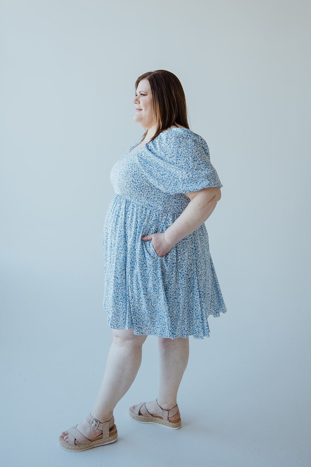 A woman is standing in profile, wearing a light blue FEMININE FLORAL PUFF SLEEVE KNEE LENGTH DRESS by Love Marlow with her hands in the dress pockets. She has shoulder-length brown hair and is wearing open-toed sandals. The background is plain white.