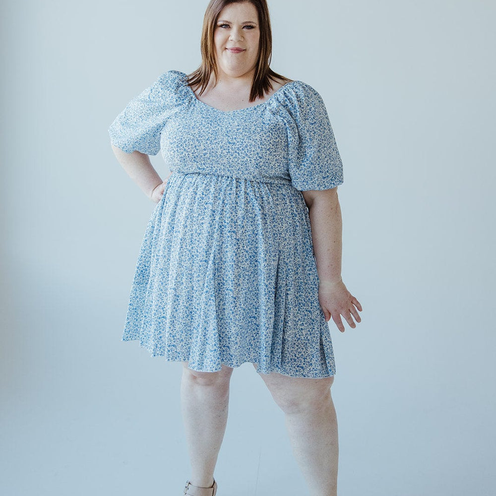 A person stands elegantly in a FEMININE FLORAL PUFF SLEEVE KNEE LENGTH DRESS by Love Marlow and beige wedge sandals against a plain white background.