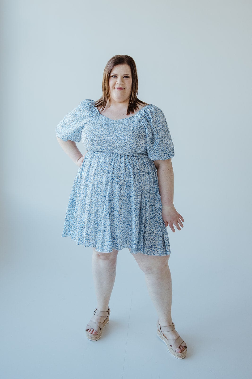 A person stands elegantly in a FEMININE FLORAL PUFF SLEEVE KNEE LENGTH DRESS by Love Marlow and beige wedge sandals against a plain white background.