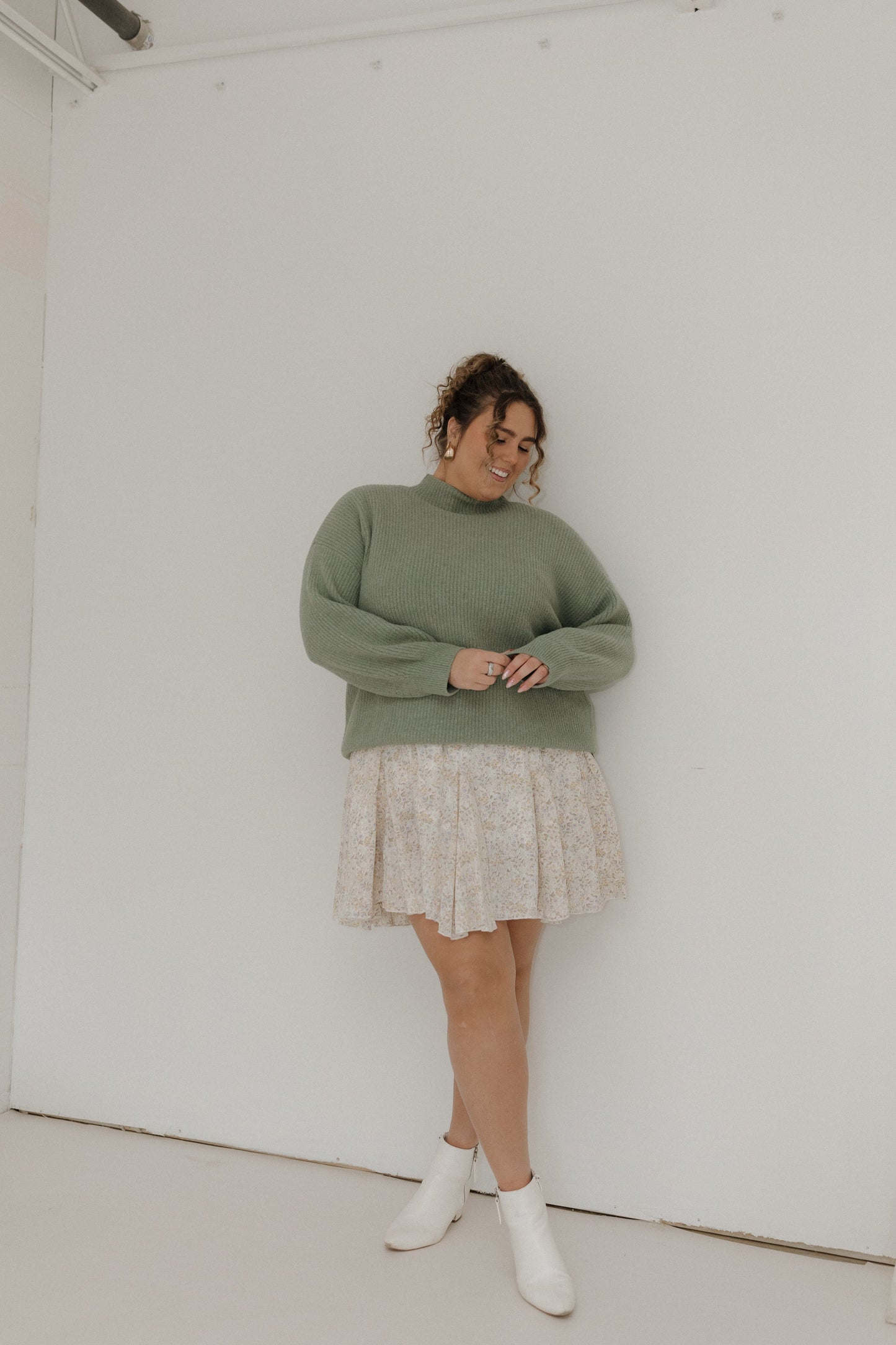 Woman wearing a green sweater and patterned skirt against a white wall