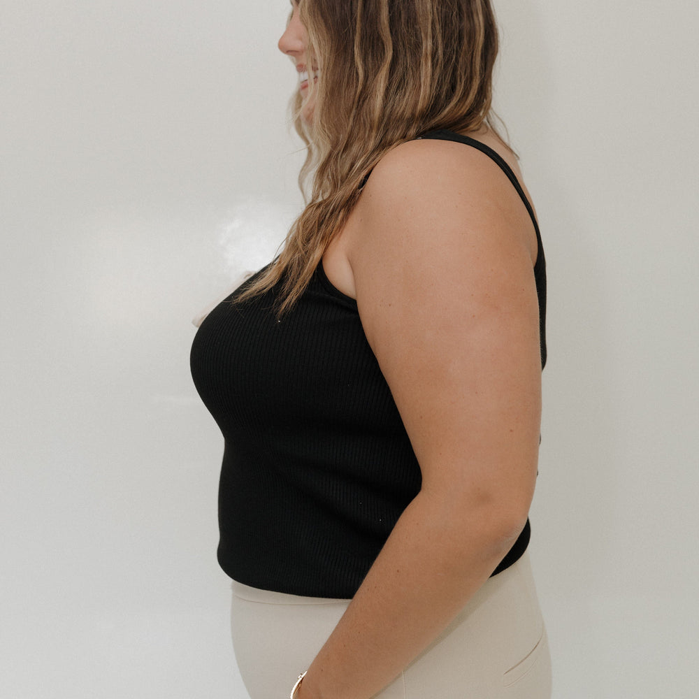 Woman wearing a black tank top and white pants against a plain background
