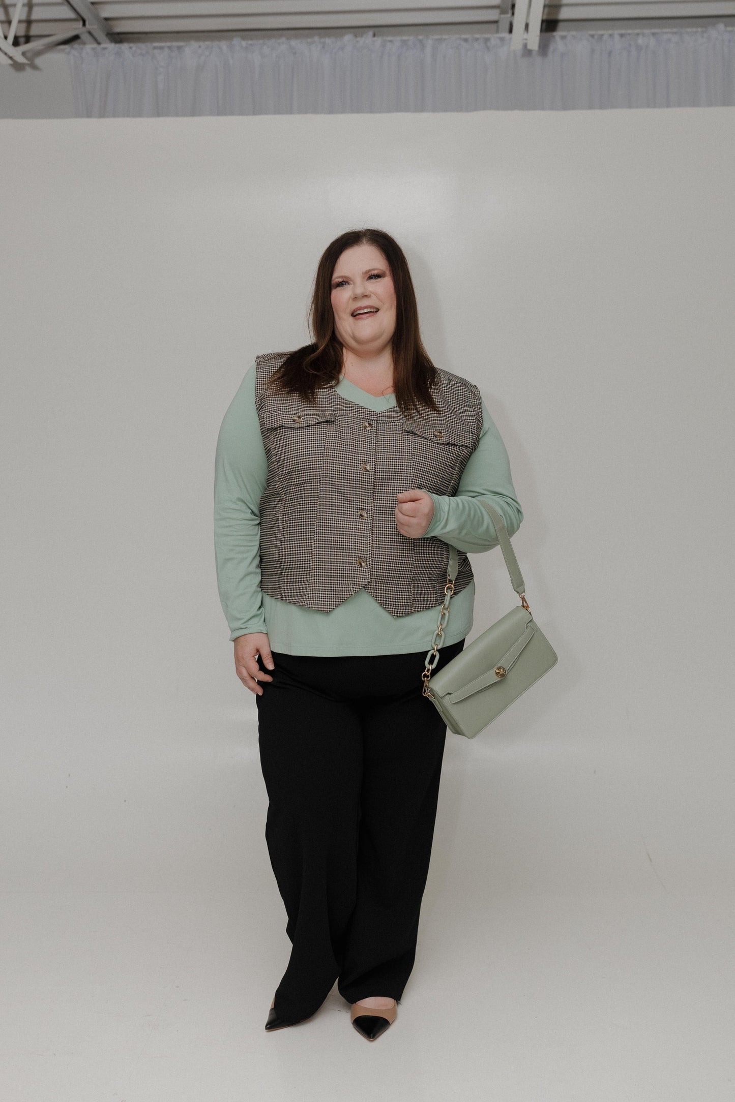 Woman wearing a green top, brown vest, black pants, and holding a light green handbag against a white background.