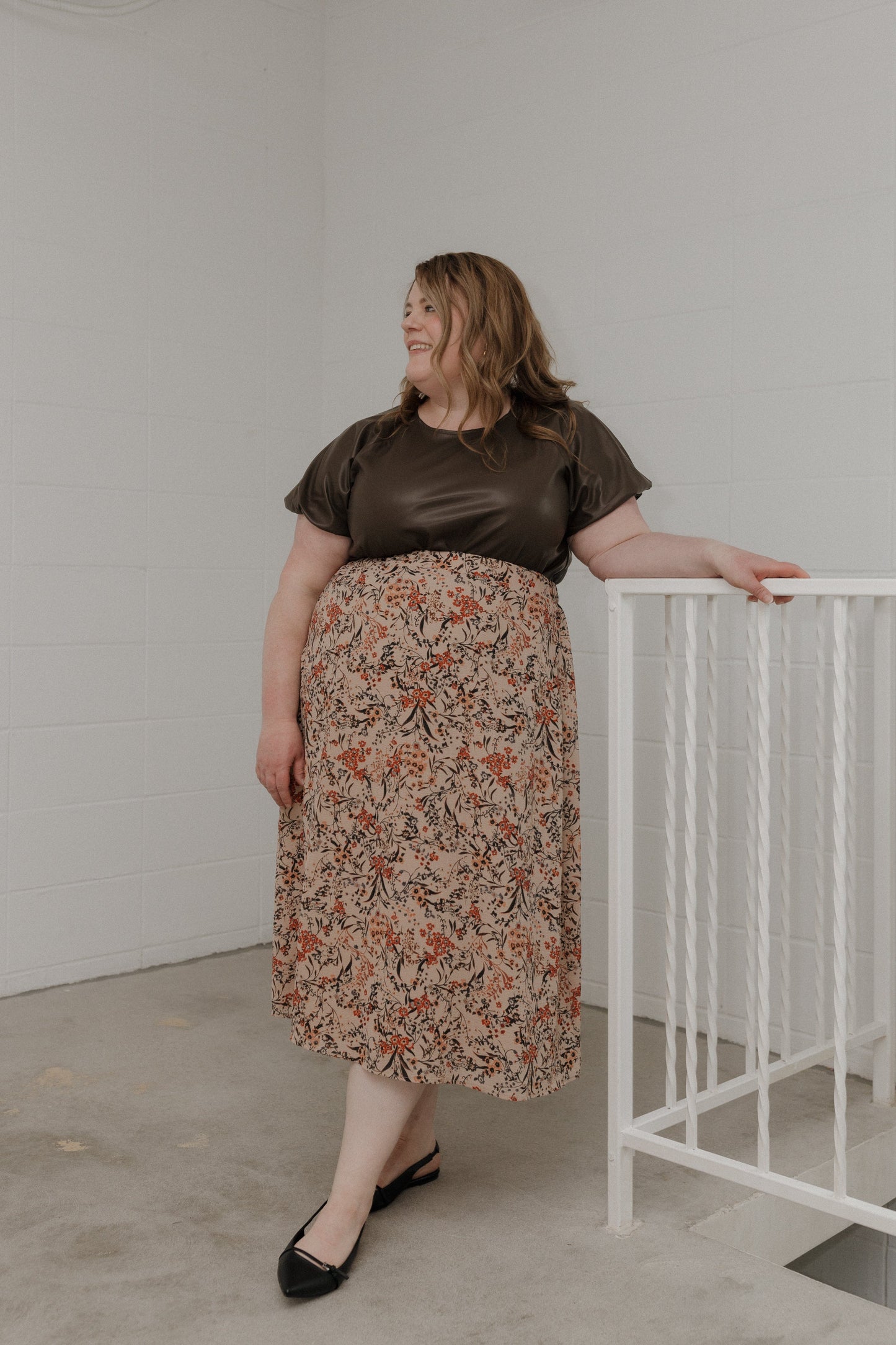 Woman wearing a brown top and floral skirt standing against a white wall.