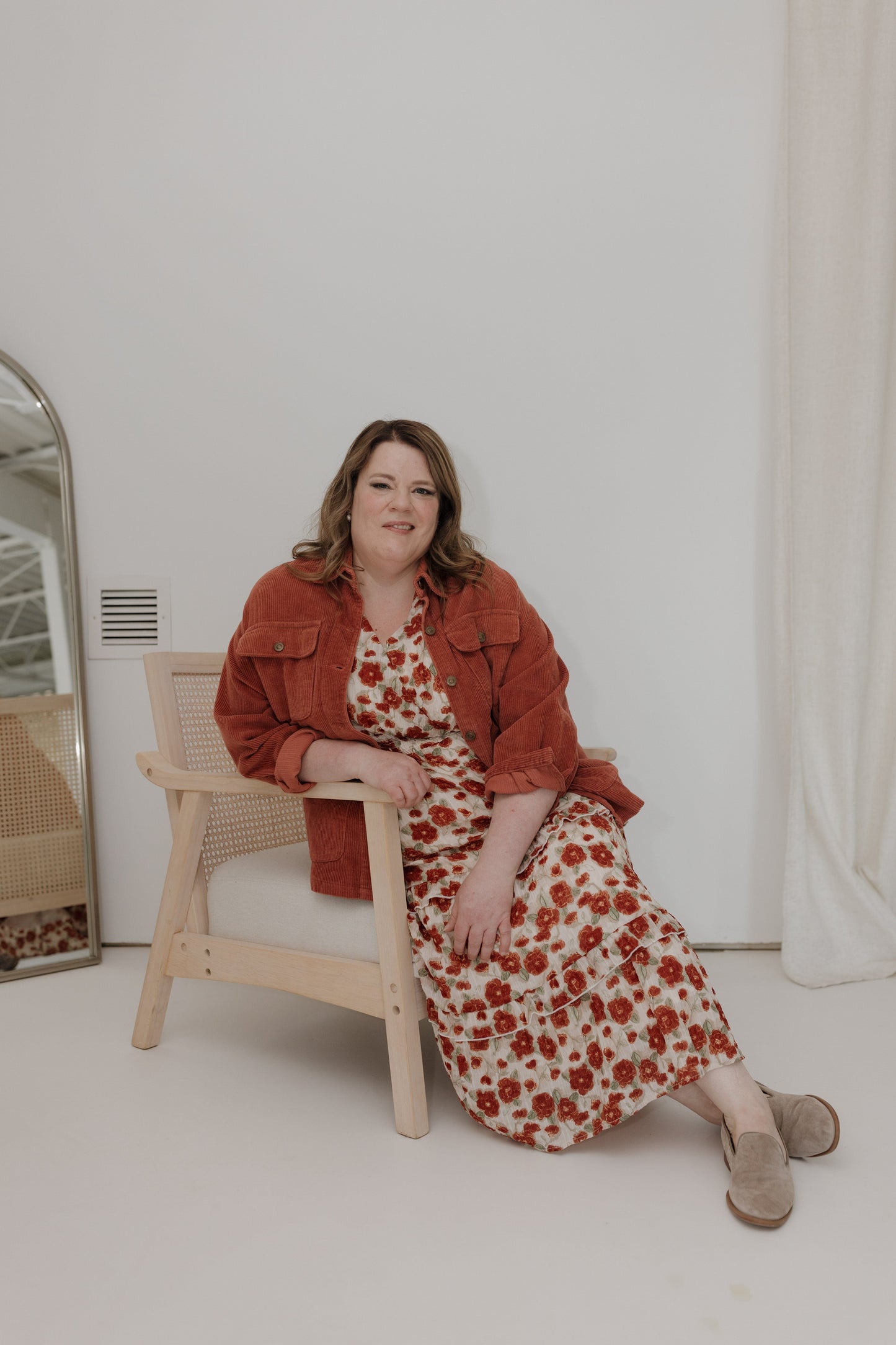 Woman in a floral dress and red jacket sitting on a chair against a white wall.