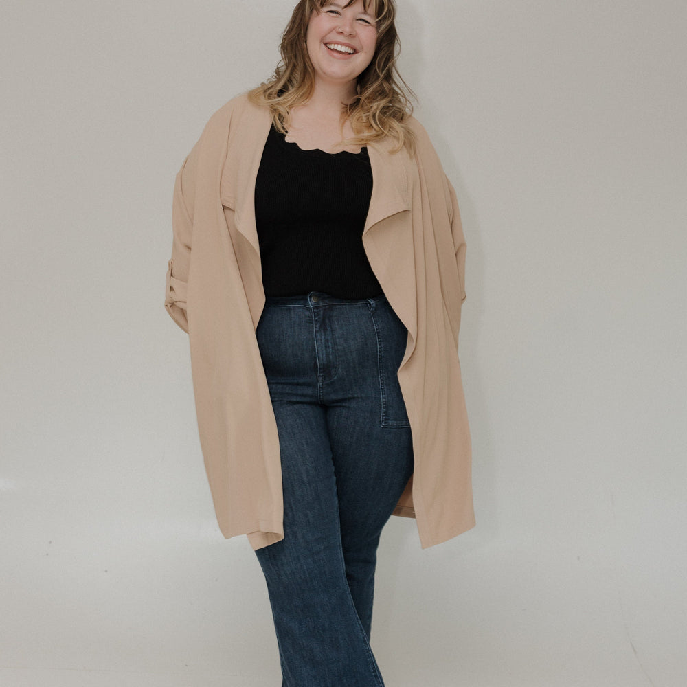 Woman wearing a beige coat, black top, and blue jeans against a plain background