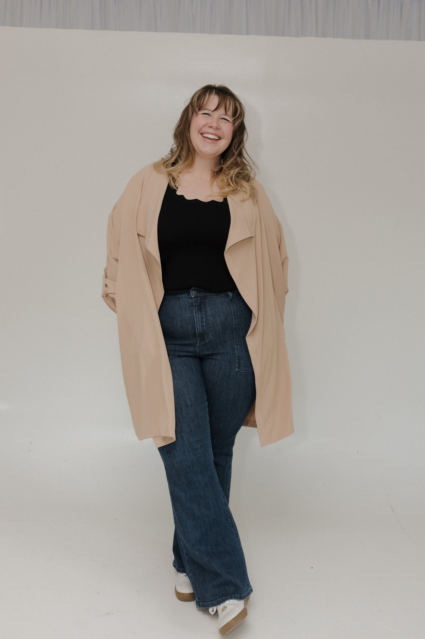 Woman wearing a beige coat, black top, and blue jeans against a plain background
