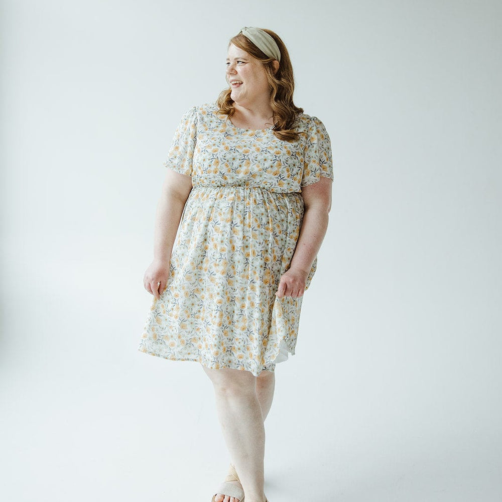 A woman in a Mikarose FLUTTER SLEEVE A-LINE KNEE LENGTH DRESS with a floral pattern and a headband stands against a plain white background, looking off to the side with a smile. She is wearing sandals and has long hair.