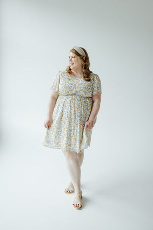 A woman in a Mikarose FLUTTER SLEEVE A-LINE KNEE LENGTH DRESS with a floral pattern and a headband stands against a plain white background, looking off to the side with a smile. She is wearing sandals and has long hair.