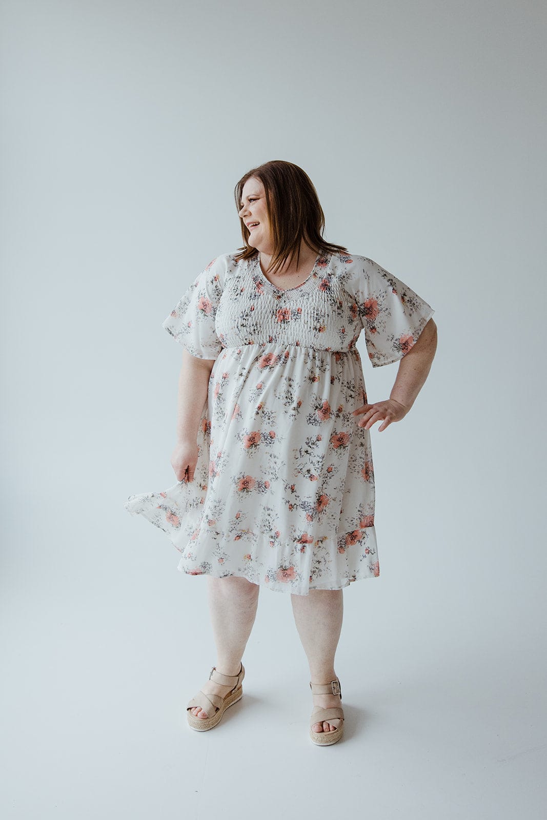 A woman in a Mikarose Flutter Sleeve Smocked V-neck Floral Dress stands against a plain background, looking to the side and smiling, with one hand on her hip and the other holding part of her dress.