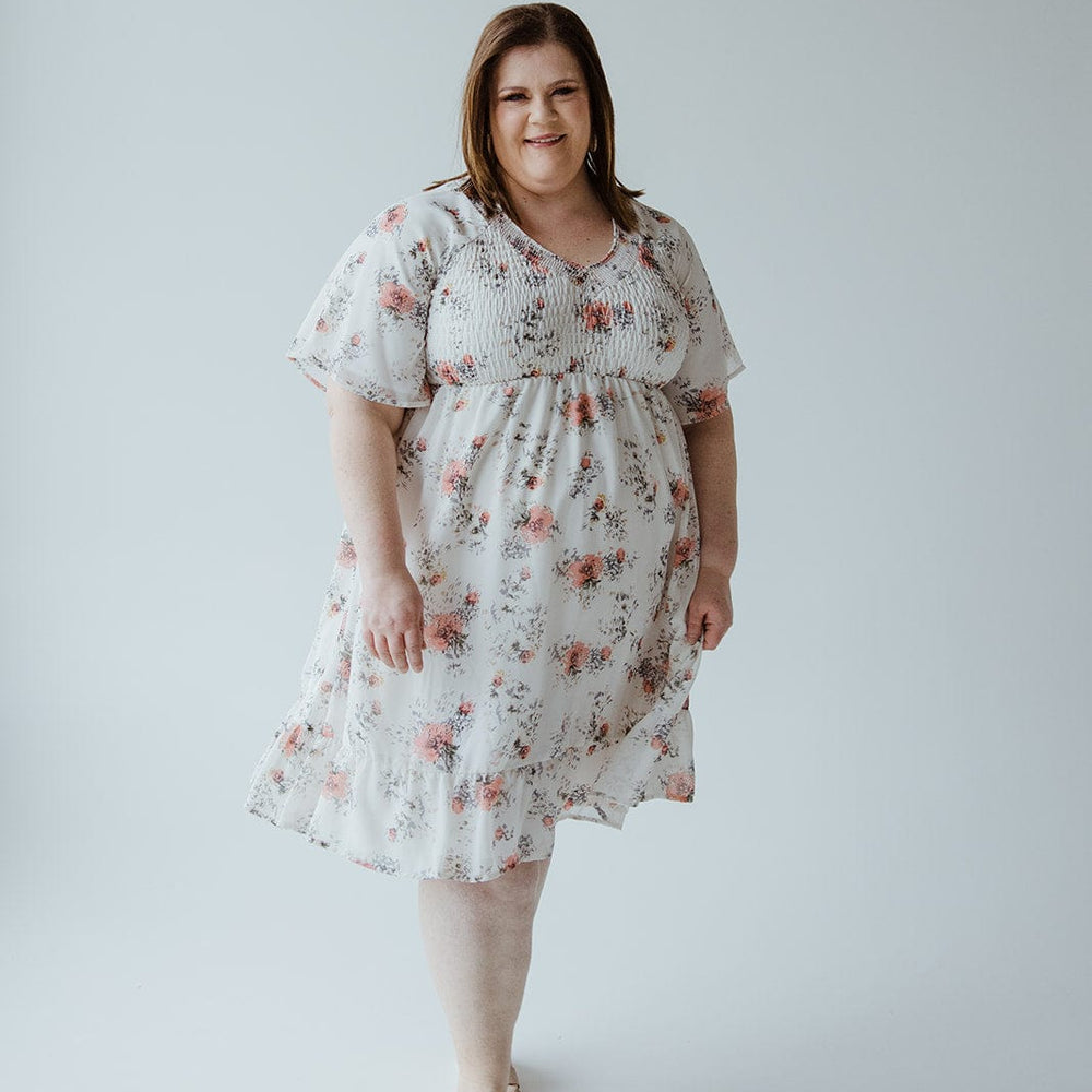 A person stands in a Mikarose Flutter Sleeve Smocked V-Neck Floral Dress paired with beige sandals against a plain background.