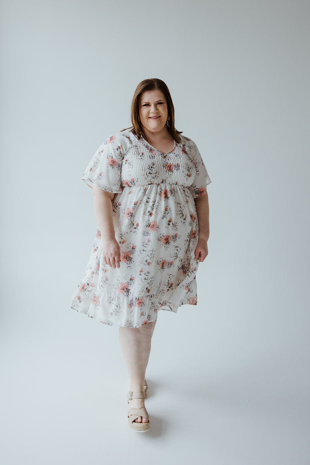 A person stands in a Mikarose Flutter Sleeve Smocked V-Neck Floral Dress paired with beige sandals against a plain background.