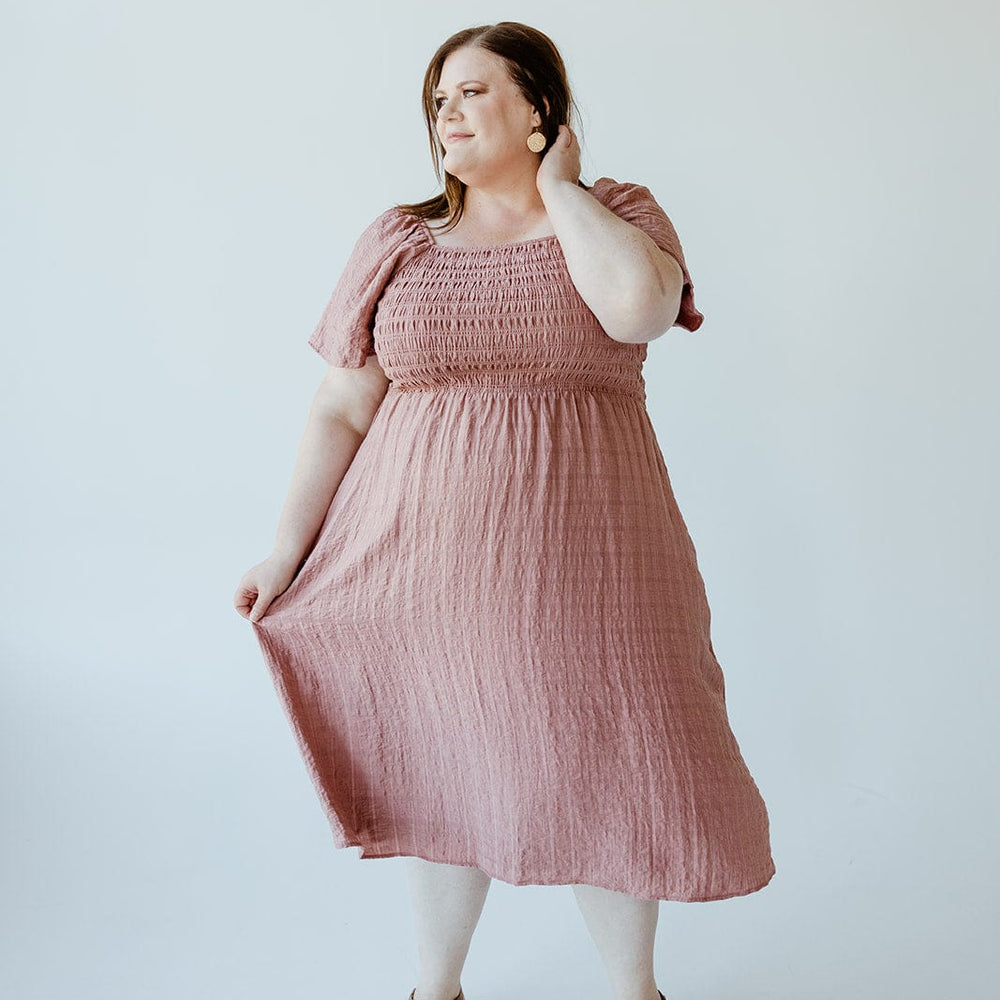 A person stands against a plain background, wearing the Mikarose FLUTTER SLEEVE TEXTURED KNEE LENGTH DRESS IN MAUVE MIST, paired with sandals, looking off to the side.