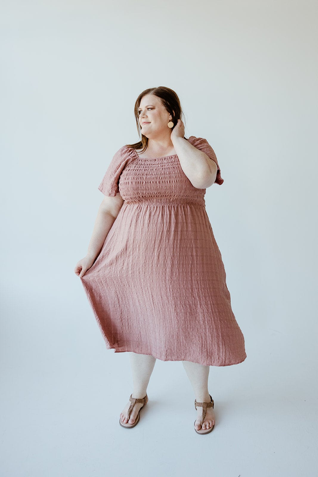 A person stands against a plain background, wearing the Mikarose FLUTTER SLEEVE TEXTURED KNEE LENGTH DRESS IN MAUVE MIST, paired with sandals, looking off to the side.