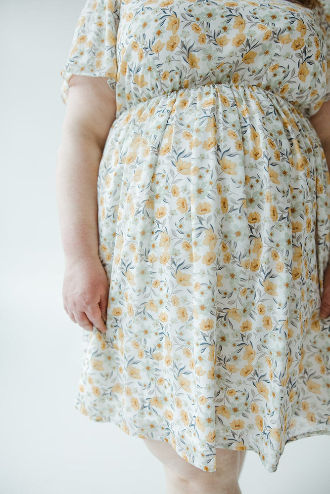 A person with long hair is standing with their back to the camera, wearing a light-colored floral FLUTTER SLEEVE A-LINE KNEE LENGTH DRESS by Mikarose and sandals.