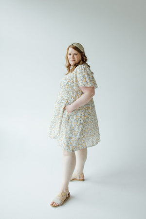 A woman stands against a plain background, wearing the FLUTTER SLEEVE A-LINE KNEE LENGTH DRESS by Mikarose and sandals. She has wavy hair and is slightly smiling.