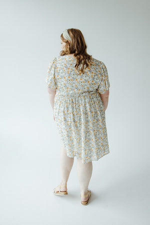 A person with long hair is standing with their back to the camera, wearing a light-colored floral FLUTTER SLEEVE A-LINE KNEE LENGTH DRESS by Mikarose and sandals.