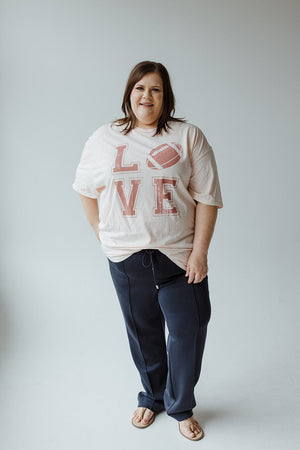 A person stands smiling in a Love Marlow "LOVE" FOOTBALL GRAPHIC TEE IN LIGHT BLUSH MINERAL WASH, paired with blue pants and flip-flops, posing against a plain background.