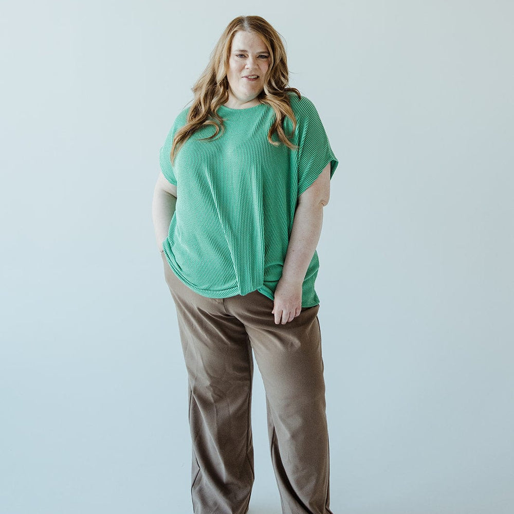 A person stands against a plain background, wearing a green top and high-waist brown pants featuring a wide-leg design, with hands in pockets.