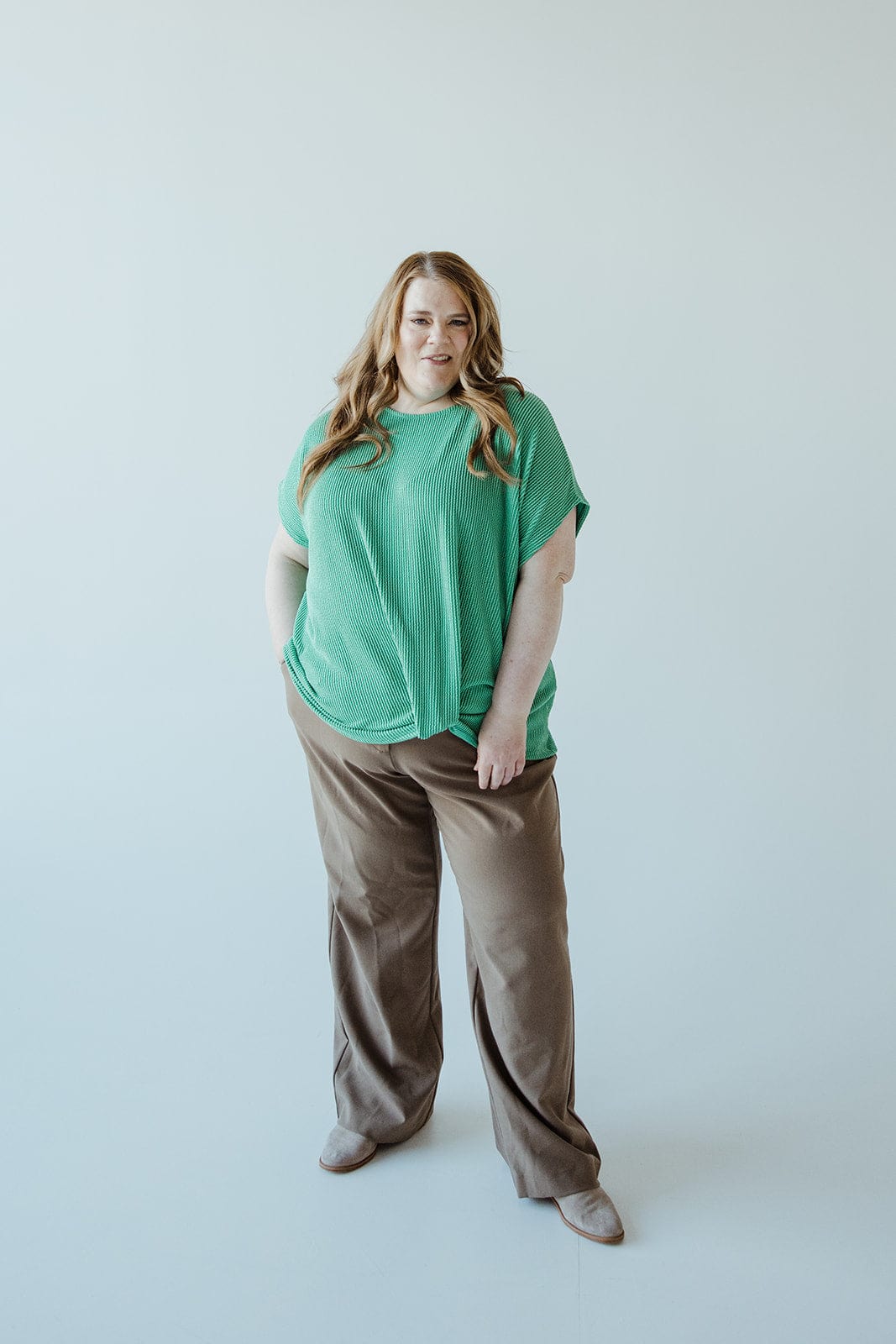 A person stands against a plain background, wearing a green top and high-waist brown pants featuring a wide-leg design, with hands in pockets.