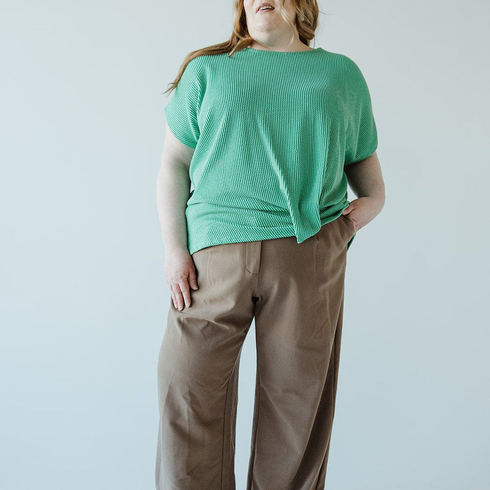 A person with long hair stands wearing a green top and stylish high-waist, brown pants featuring a chic wide-leg design against a plain background.