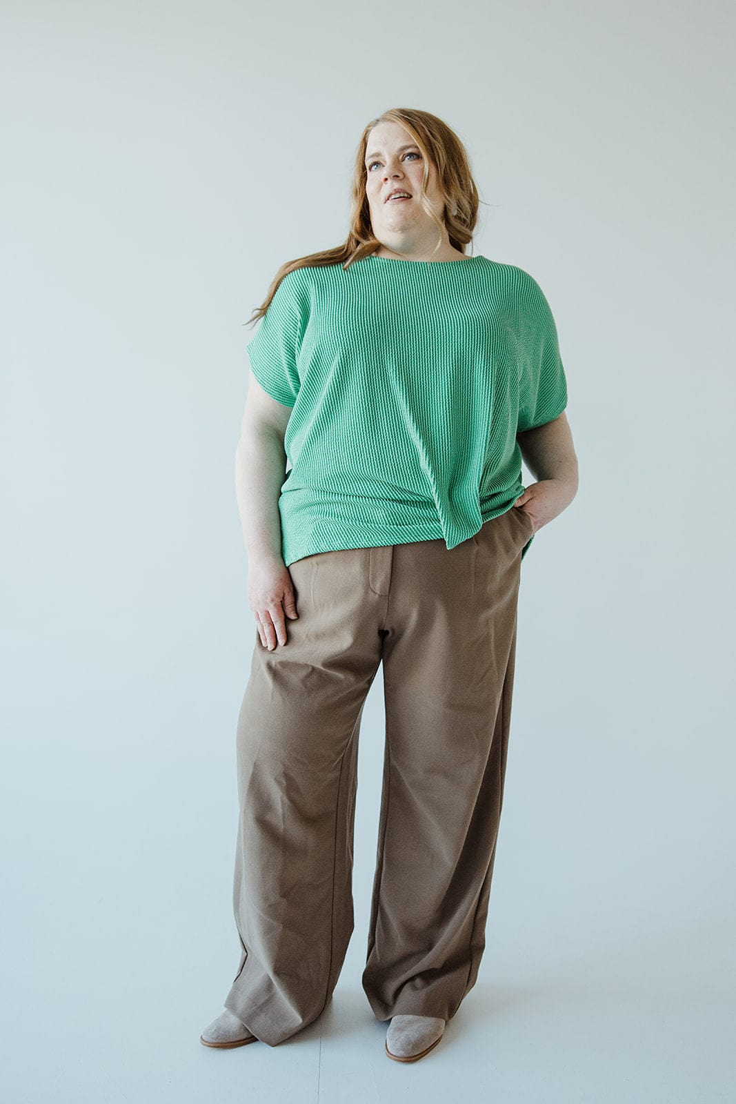 A person with long hair stands wearing a green top and stylish high-waist, brown pants featuring a chic wide-leg design against a plain background.