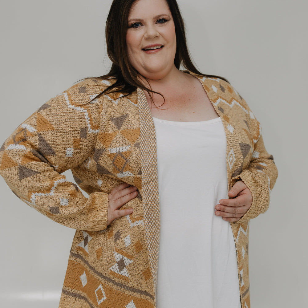 Woman wearing a patterned beige cardigan over a white top and brown pants on a plain background