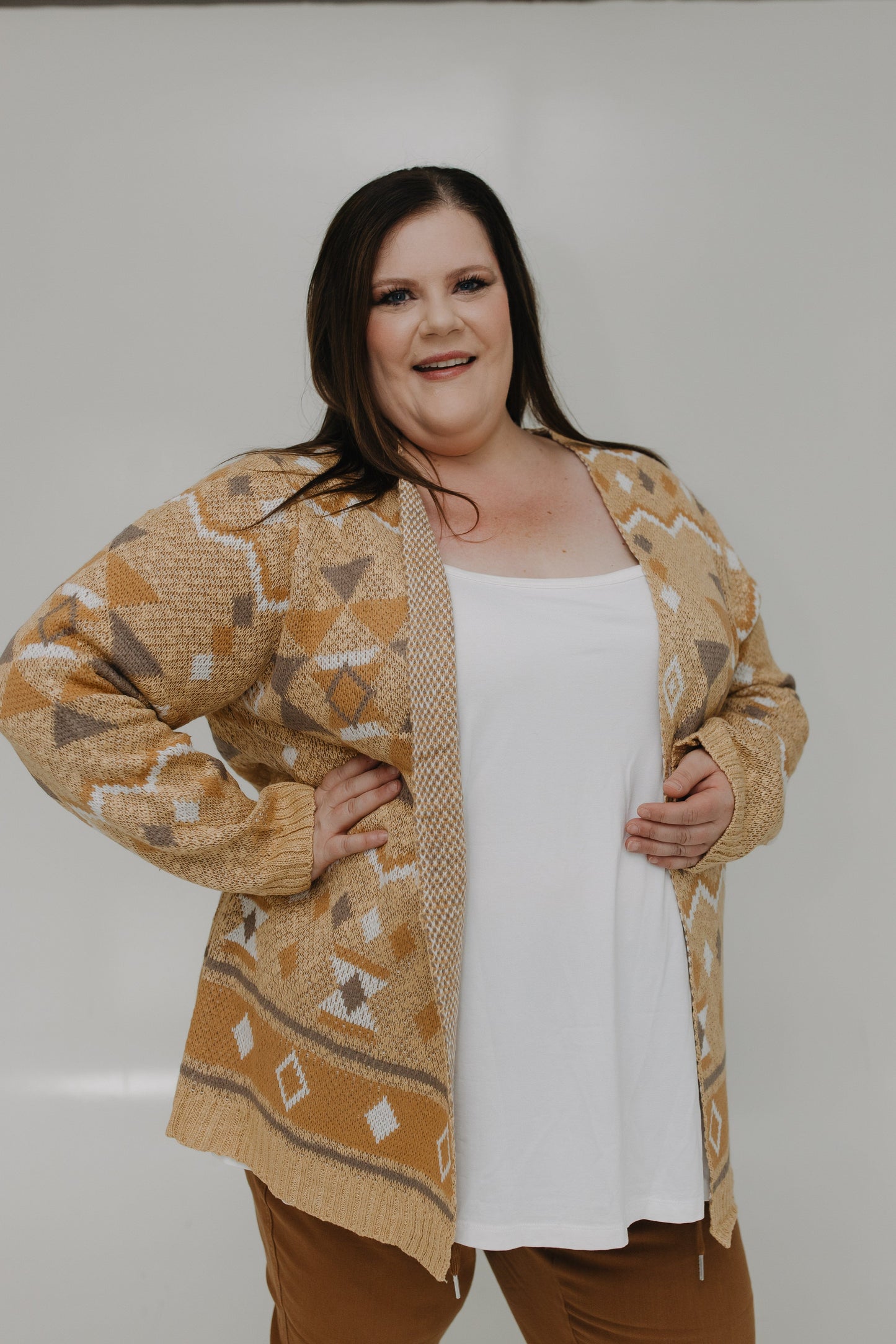 Woman wearing a patterned beige cardigan over a white top and brown pants on a plain background