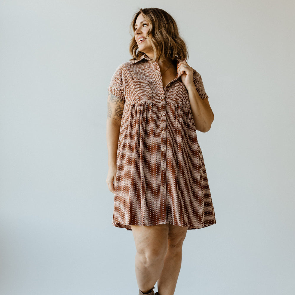 A person wearing a knee-length geometric-patterned dress and brown ankle boots stands on a light gray background, looking to the side with a slight smile.