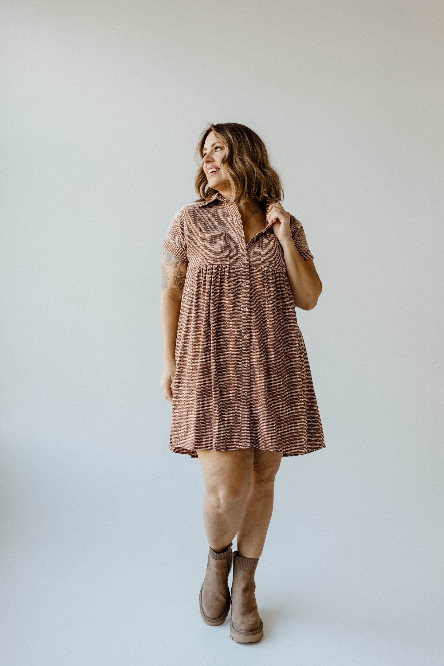 A person wearing a knee-length geometric-patterned dress and brown ankle boots stands on a light gray background, looking to the side with a slight smile.