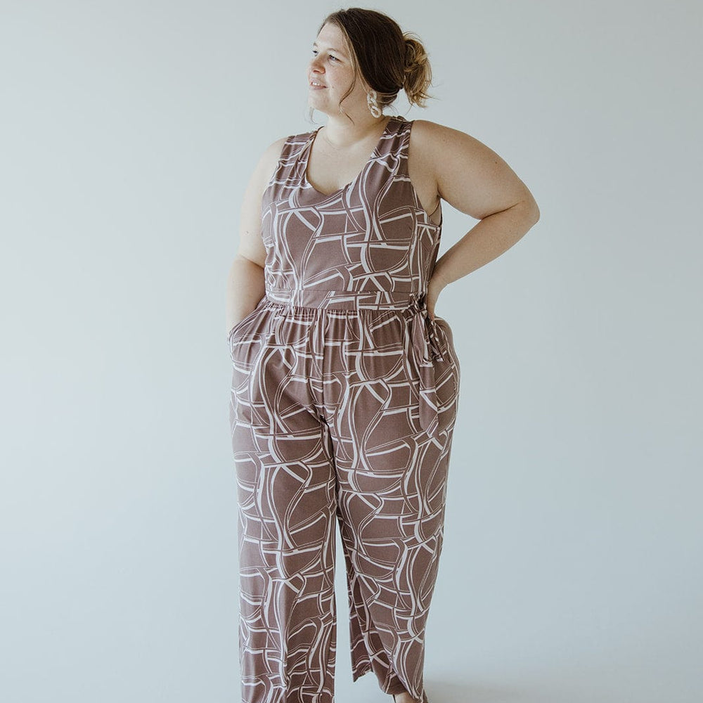 A person stands against a plain background, wearing a brown and white Geometric V-Neck Jumpsuit with an elastic waist and hands in the pockets, looking to the side. The jumpsuit is from the brand Gilli and includes a tie.