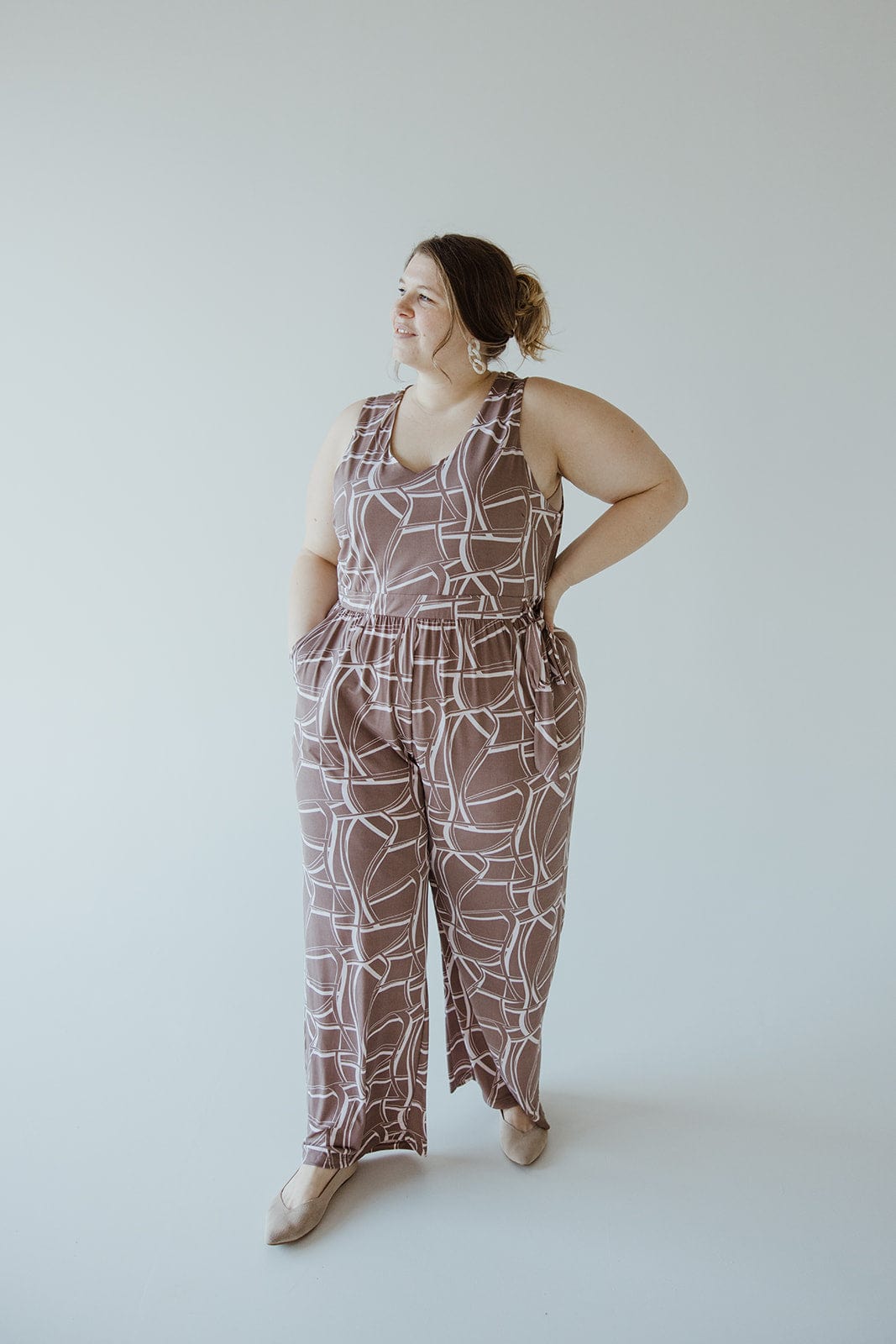 A person stands against a plain background, wearing a brown and white Geometric V-Neck Jumpsuit with an elastic waist and hands in the pockets, looking to the side. The jumpsuit is from the brand Gilli and includes a tie.