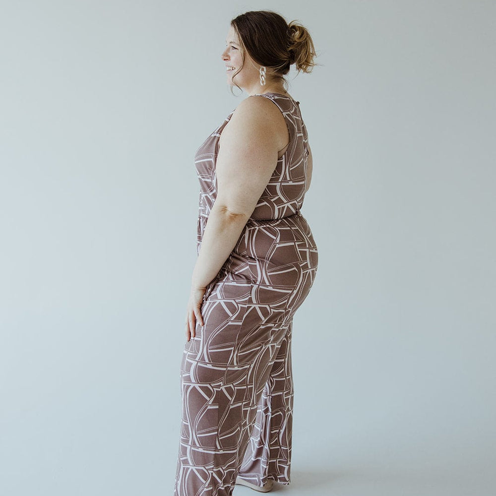 A woman stands in profile against a plain background, wearing the Gilli Geometric V-Neck Jumpsuit with Tie and beige heels. She has her hair tied back and is smiling.