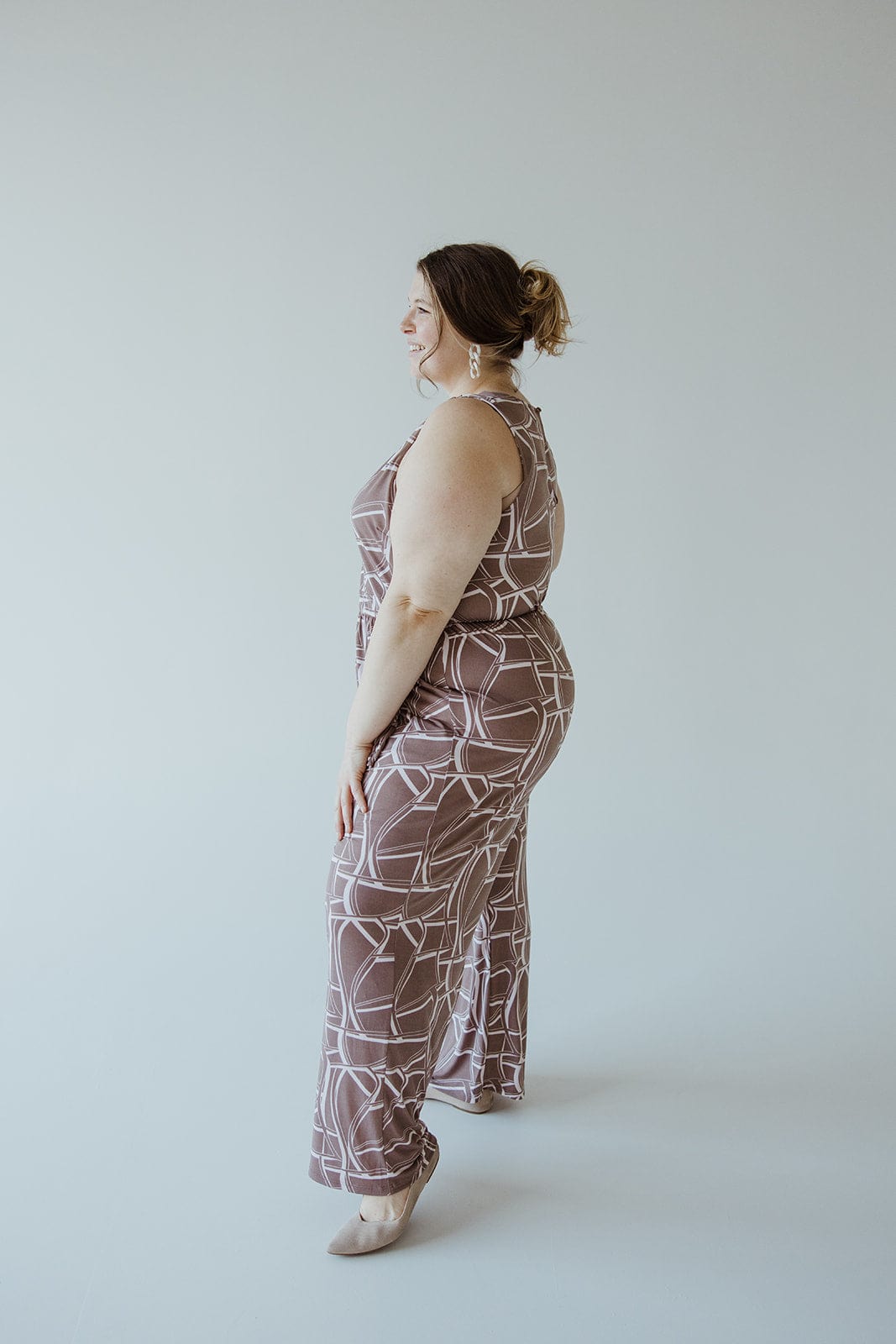 A woman stands in profile against a plain background, wearing the Gilli Geometric V-Neck Jumpsuit with Tie and beige heels. She has her hair tied back and is smiling.