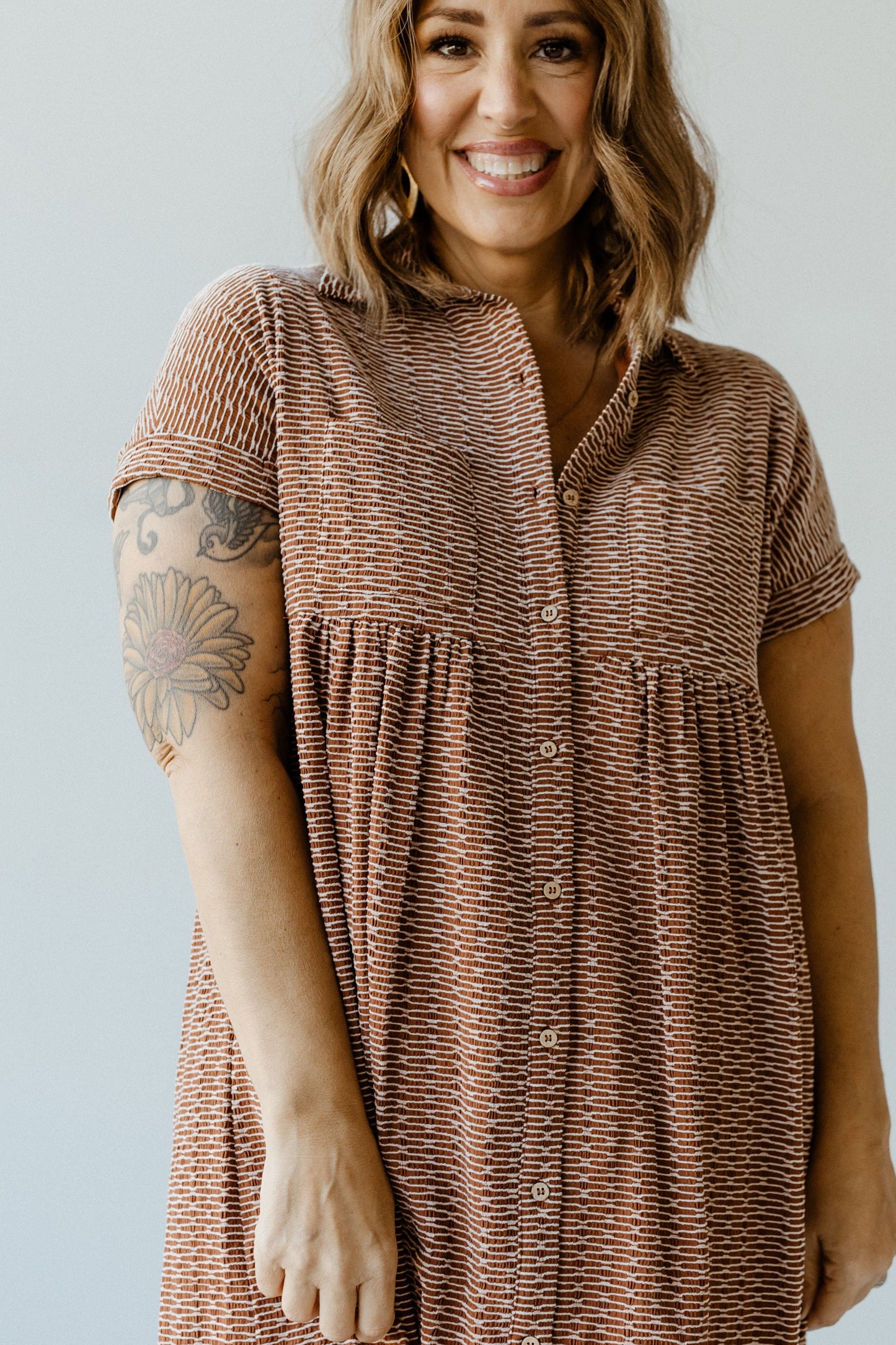 A person with shoulder-length hair and a sunflower tattoo on their arm wears a knee-length dress with a textured pattern, standing against a plain background.