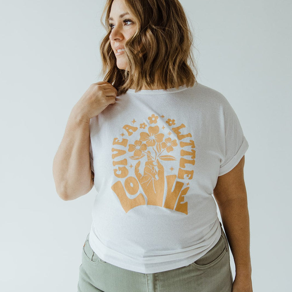 A person with shoulder-length hair is wearing a white graphic tee featuring "Give a Little Love" and floral print, paired with green pants that have a rolled cuff. They're standing in front of a plain background.