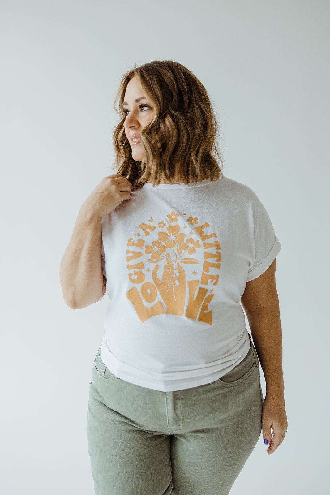 A person with shoulder-length hair is wearing a white graphic tee featuring "Give a Little Love" and floral print, paired with green pants that have a rolled cuff. They're standing in front of a plain background.